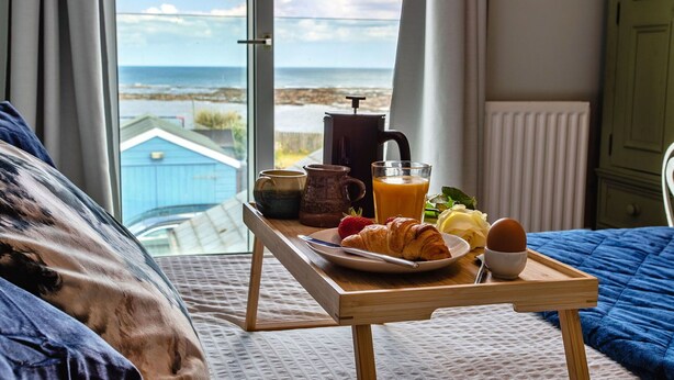 A bedroom with a view of the sea, a bedside table with breakfast items, and a blue bedspread.
