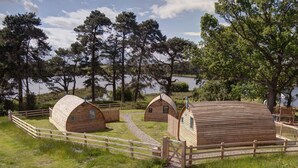 Una hilera de cabañas de madera con techos a dos aguas, rodeadas por una valla de madera, con un paisaje de árboles y una masa de agua de fondo.
