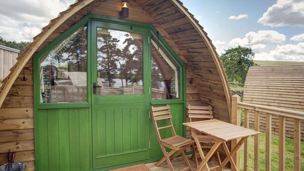 Una cabaña de madera con puerta verde, una pequeña mesa y silla, y vistas del paisaje circundante.