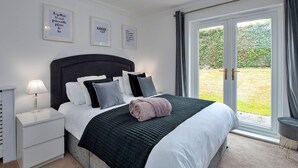 A bedroom with a large bed, a nightstand, a lamp, and a window with a view of a garden. - Lovely apartment for 5 guests with pool, WIFI, TV and balcony (Tenby)