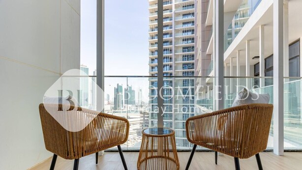 A modern apartment with large windows offering a city view, two wicker chairs, and a small round table. - Bloomfields Skyline 1Br In Al Maryah  (Abu Dhabi)