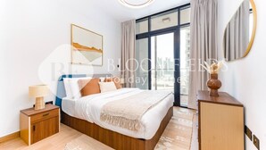 A modern bedroom with a large bed, a bedside table, a nightstand, a mirror, and a view of the city through large windows. - Bloomfields Skyline 1Br In Al Maryah  (Abu Dhabi)