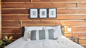 A wooden-paneled wall with three framed designs, a bed with white bedding and striped pillows, and a bedside table with a lamp.