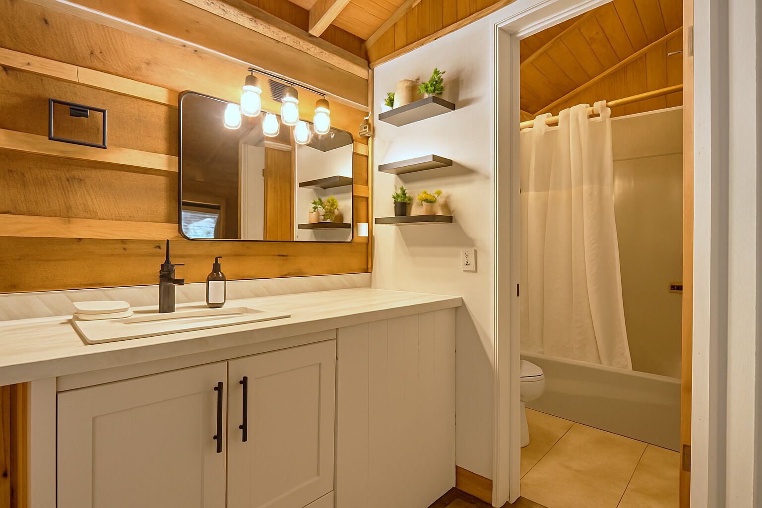A bathroom with a wooden ceiling, a large mirror with lights, a sink, cabinets, and a shower area.
