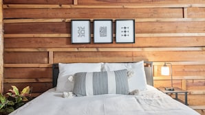 Pared de madera con tres diseños enmarcados, una cama con ropa de cama blanca y almohadas a rayas, y una mesita de noche con una lámpara.