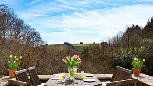 An outdoor dining area with a table set for four, surrounded by chairs with striped cushions. There are potted plants with yellow flowers on either side of the table.