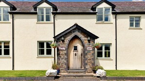 A farmhouse-style building with a central wooden door, stone arch, and hanging flower baskets.