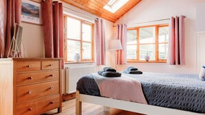 A cozy bedroom with a wooden ceiling, two windows with curtains, a bed with a blanket, a dresser, and a lamp.