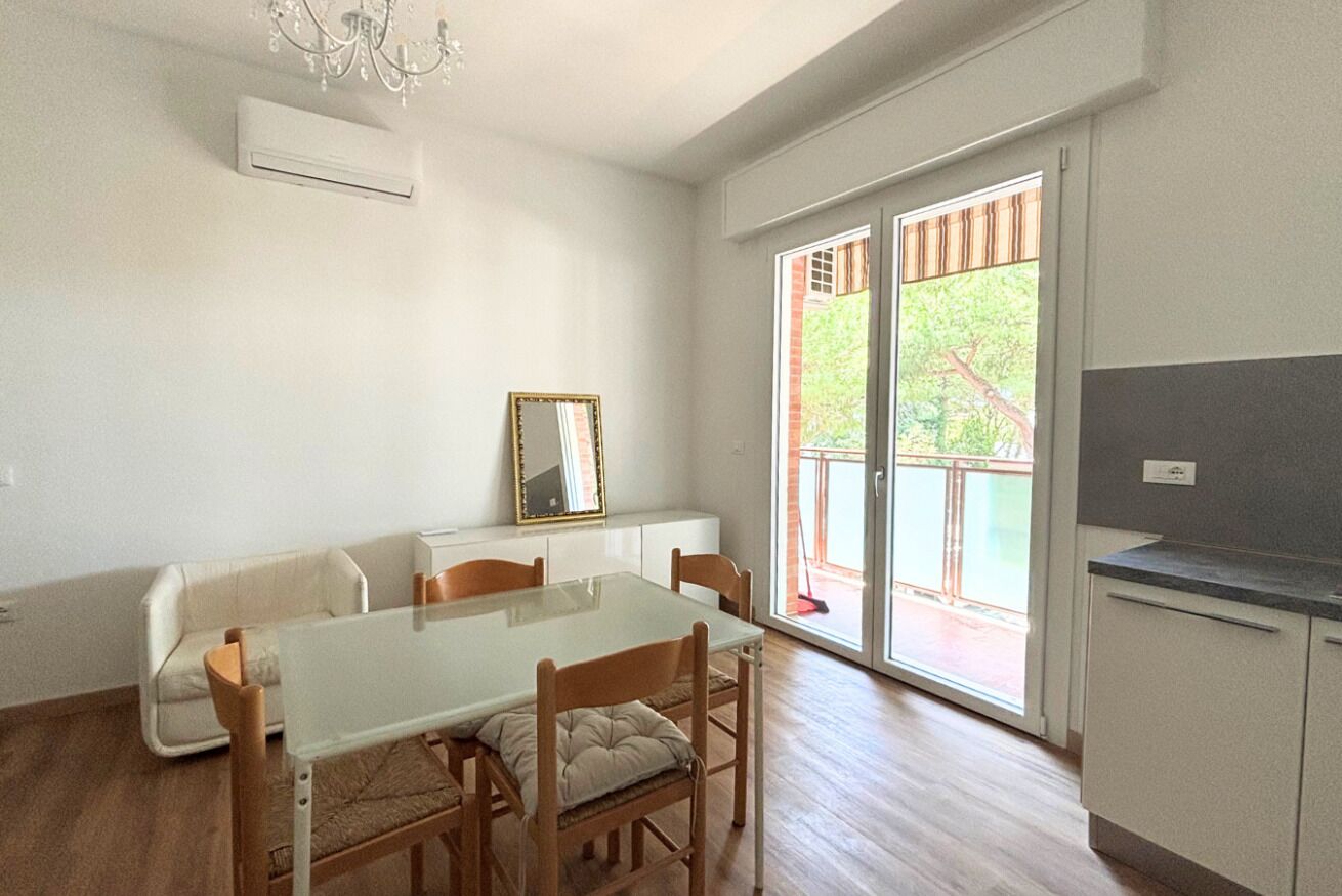 Zona pranzo con tavolo in vetro, sedie in legno, divano bianco, uno specchio e una porta vetrata scorrevole che dà su un’area esterna.