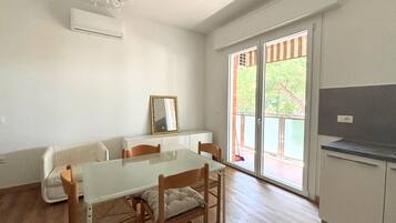 Zona pranzo con tavolo in vetro, sedie in legno, divano bianco, uno specchio e una porta vetrata scorrevole che dà su un’area esterna.