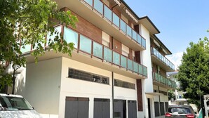Un immeuble résidentiel à plusieurs étages, doté d’un garage, d’un balcon à l’étage supérieur et d’une voiture garée devant.