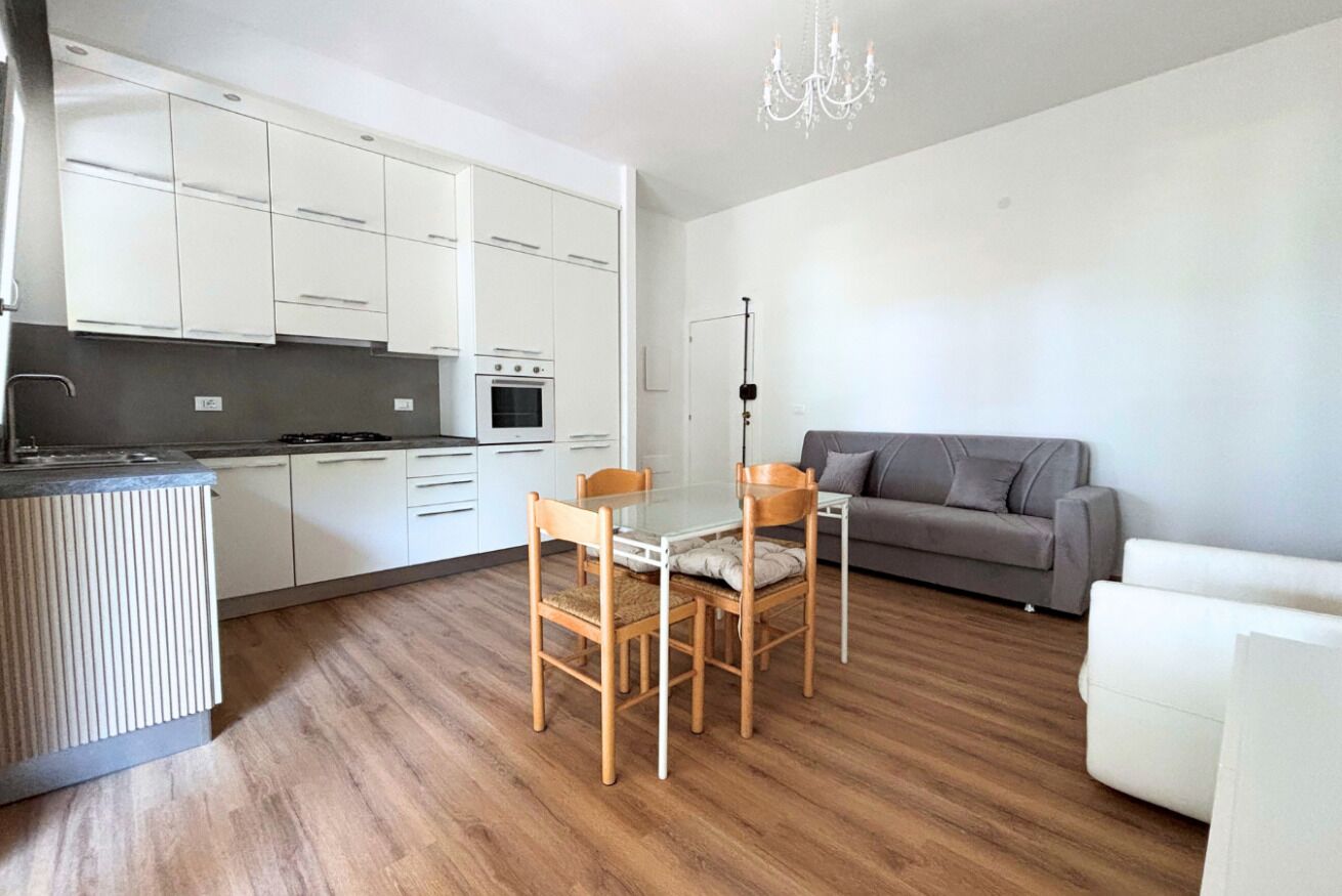 Una cucina compatta con mobili bianchi, un tavolo da pranzo con sedie, un divano grigio e un lampadario.