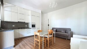 Une cuisine compacte avec des meubles blancs, une table à manger avec des chaises, un canapé gris et un lustre.
