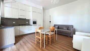 Una cucina compatta con mobili bianchi, un tavolo da pranzo con sedie, un divano grigio e un lampadario.