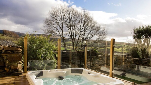 Un jacuzzi en una terraza de madera con una barandilla de vidrio, con vistas a un paisaje rural con árboles y colinas.