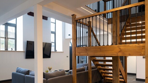 A spacious living area with a wooden staircase, a grey sofa, and a flat-screen TV mounted on the wall.