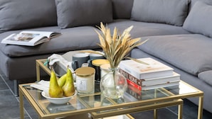 A modern living room with a glass coffee table, a bowl of pears, a scented candle, a vase with dried plants, and a stack of books.