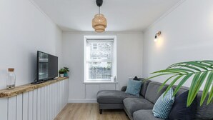 Un espacio habitable pequeño con un sofá gris, un televisor sobre una estantería de madera, un ventanal con persianas y una lámpara colgante.