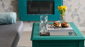 Une table basse de couleur turquoise avec un plateau en verre, un verre transparent, un livre et deux pâtisseries.