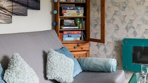 Un salon avec un canapé gris, des coussins bleus et blancs, une bibliothèque en bois remplie de livres et de jeux de société, et un meuble de rangement turquoise contenant des verres.