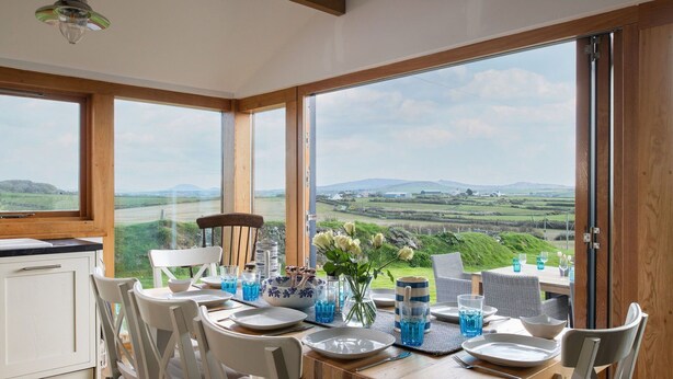 A dining area with a table set for four, large windows offering a view of the countryside, and a wooden ceiling. - Stunning  villa for 8 guests with WIFI, TV and balcony (Aberdaron)