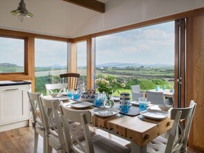 Uma sala de jantar com uma mesa posta para quatro pessoas, grandes janelas com vista para o campo, e teto de madeira.