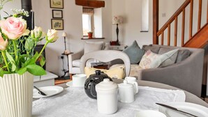 Una sala de estar acogedora con una mesa redonda dispuesta para el té, con teteras y tazas blancas, un florero con flores, y un sofá gris con cojines.