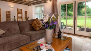 Un salon avec un canapé marron, une table basse avec des magazines, un vase de fleurs et une vue sur un jardin, visible à travers une porte vitrée.