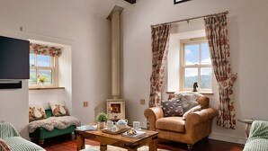 Una sala de estar acogedora con chimenea, sofás tapizados en verde, una mesa de centro de madera y cortinas con estampado floral.