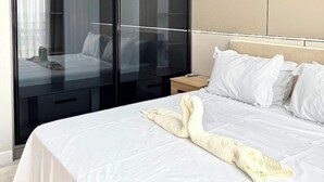 A modern hotel room with a large bed, white bedding, a black wardrobe with glass doors, and a neutral color scheme.
