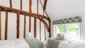 A room with two beds, wooden beams on the ceiling, a window with a patterned valance, and a view of greenery outside. - Tuffs Farm (Kings Langley)