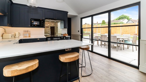 A modern kitchen with dark cabinetry, a large island with stools, and a view of an outdoor patio through large sliding doors. - Tuffs Farm (Kings Langley)