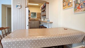 Eine kleine Küche mit Kühlschrank, Spüle und Holzschränken. Im Vordergrund steht ein Esstisch mit einem Tischlein in Punkte.