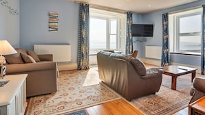 A living room with a sofa, a coffee table, and a TV. There are curtains and a window with a view.