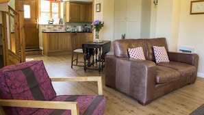 Una sala de estar acogedora con un sofá de cuero marrón, un sillón estampado y una mesa de comedor con un jarrón de flores. Se ve una cocina con armarios de madera y una escalera al fondo.