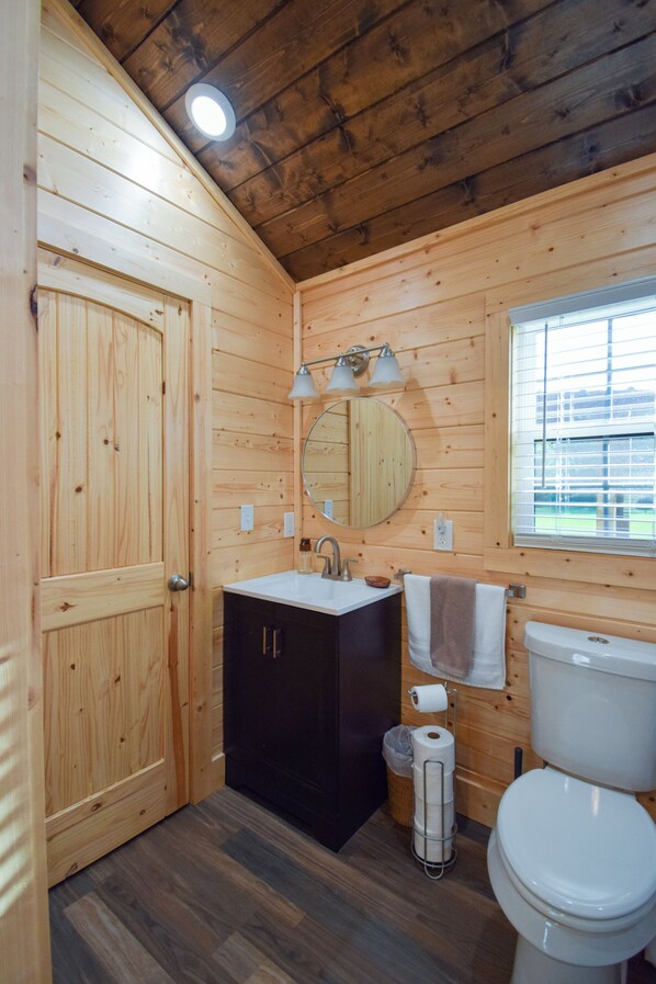 A wooden cabin bathroom with a sink, mirror, toilet, and a window with blinds. - Welcome to Cabin by the Meadow-your perfect retreat on the outskirts of town! (Texarkana)