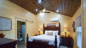 Un dormitorio de cabaña de madera con una cama de madera, dos mesitas de noche con lámparas, un ventilador de techo y una ventana con persianas.