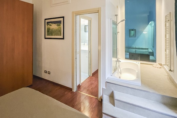 A bedroom with a wooden floor, a bed, a door leading to another room, and a bathroom with a bathtub and shower.