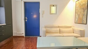 Ett rum med en blå dörr, en beige soffa, ett soffbord i glas, ett element och ett litet fönster.