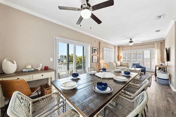 A dining area with a long table, wicker chairs, ceiling fans, and a view through large windows.