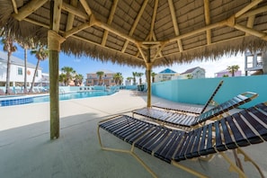 A pool area with lounge chairs, a thatched roof, and a view of buildings in the background.