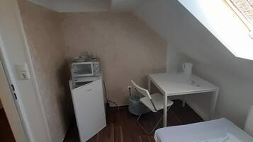 A small, compact room with a sloped ceiling, a bed, a desk, a chair, and a microwave.