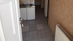 A narrow hallway with a kitchen area featuring a sink, stove, and refrigerator. A radiator is visible on the right, and the walls are plain with a beige accent. - 2-Rent Group Hostel Rooms&Apartments EVE168 (Munich)