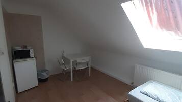 Un apartamento pequeño de una sola habitación con techo inclinado, tragaluz, cama, mesita, silla, microondas y radiador.