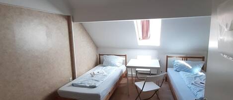 A small, single-room accommodation with two beds, a desk, and a chair.