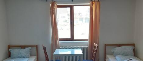 A room with two single beds, a table with chairs, a radiator, and a window with curtains.