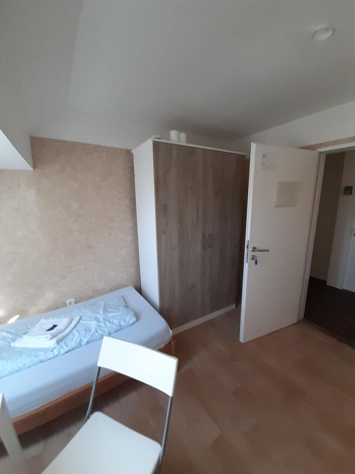 A bedroom with a bed, a wooden wardrobe, a white chair, and a door leading to another room.