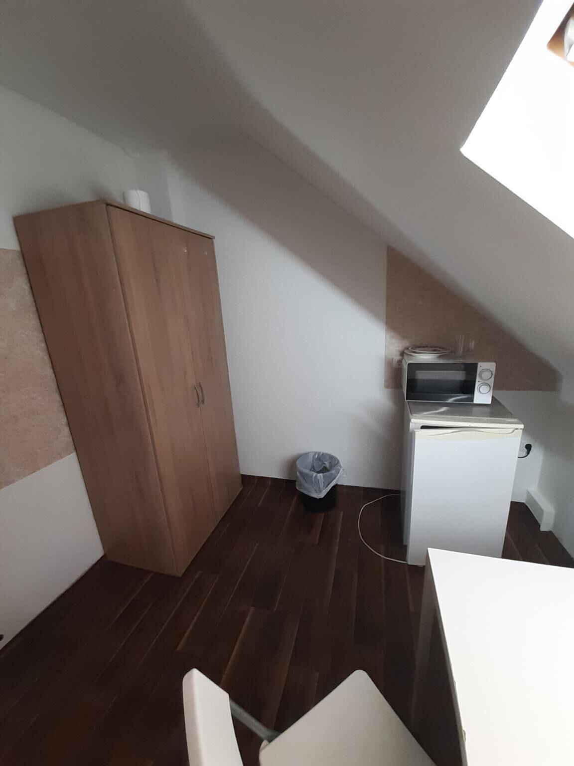 Une petite pièce étroite, avec une armoire en bois, un petit appareil électroménager blanc et une poubelle.