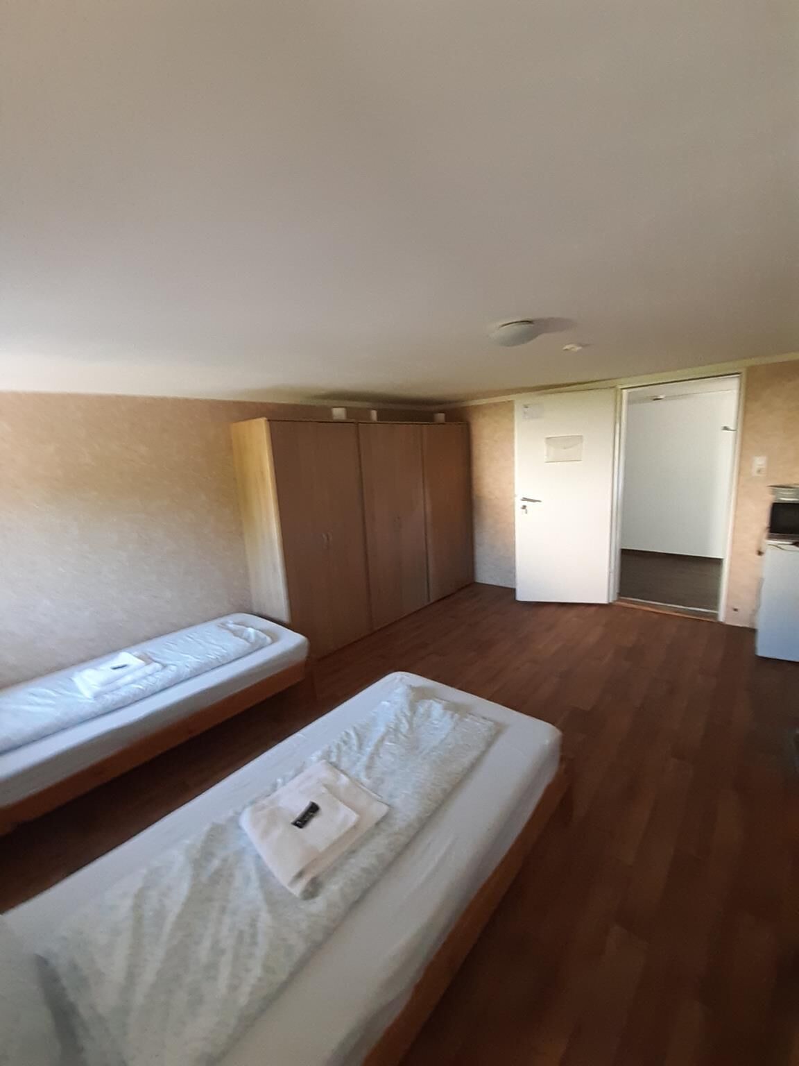 Una habitación con dos camas individuales, suelo de madera, un armario y una puerta que da a otra habitación.