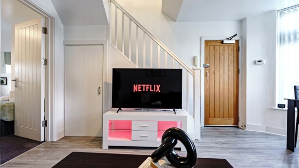 Una sala de estar con televisor mostrando 'NETFLIX', una escalera y una puerta de madera.
